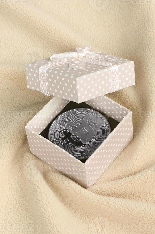 The silver bitcoin lies in a small orange gift box with a small bow on a blanket made of soft and fluffy light orange fleece fabric with a large number of relief folds photo