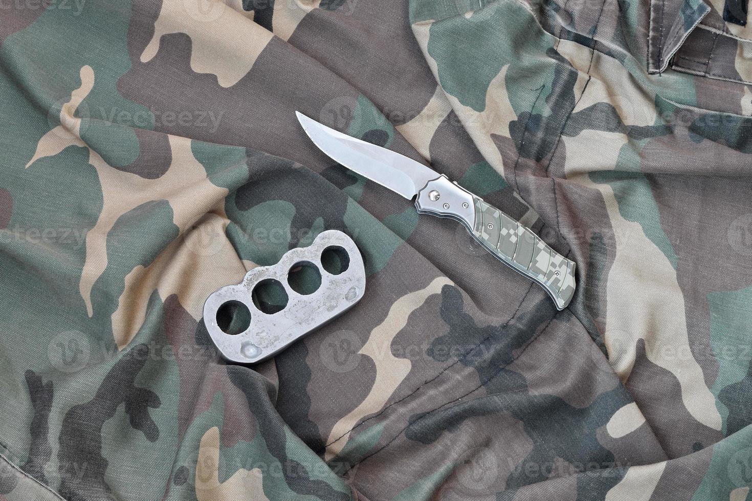 cuchillo y nudillos de latón sobre un fondo de ropa de camuflaje. herramientas para la autodefensa o ataques de delincuentes foto