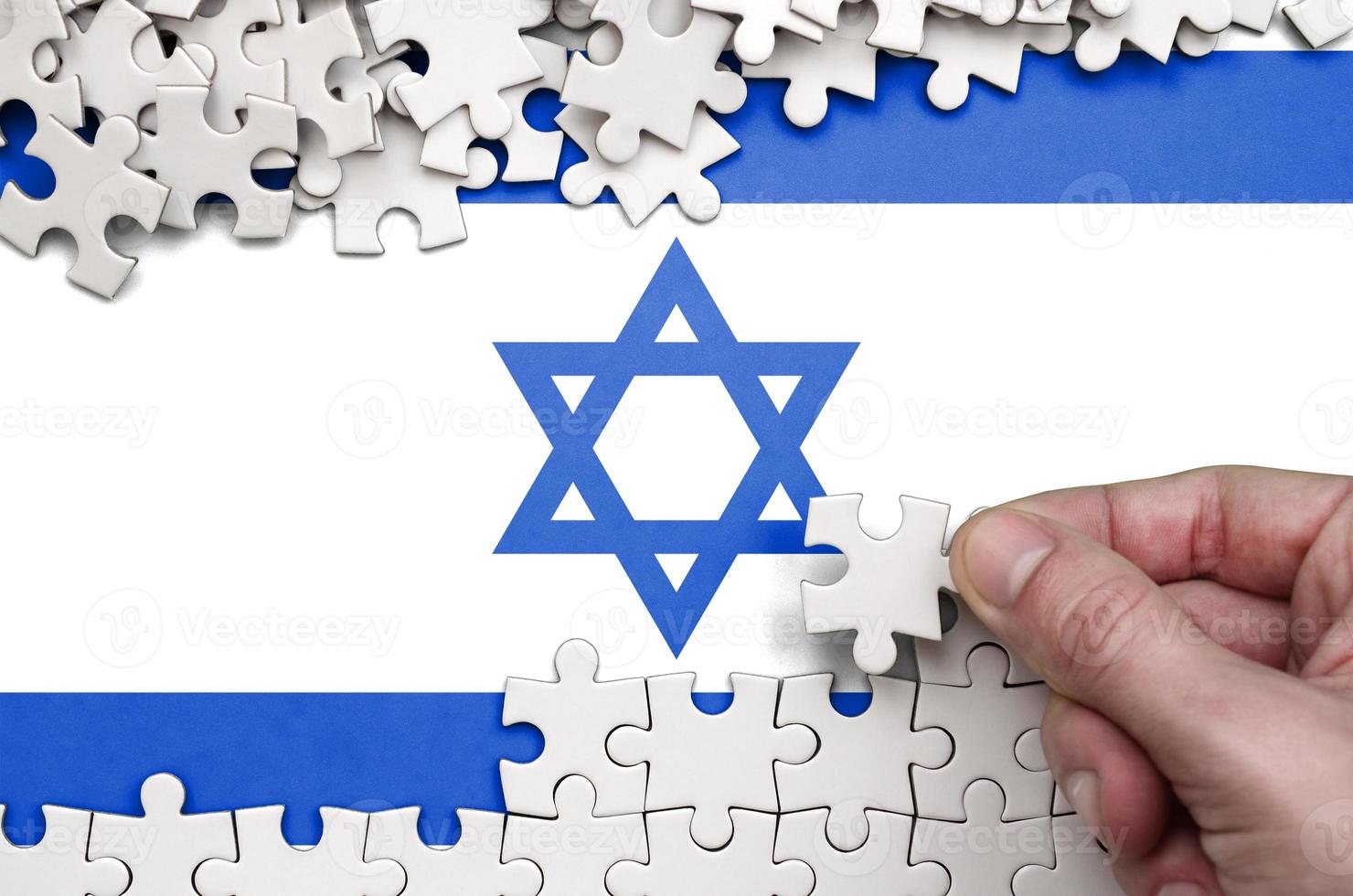 Israel flag is depicted on a table on which the human hand folds a puzzle of white color photo