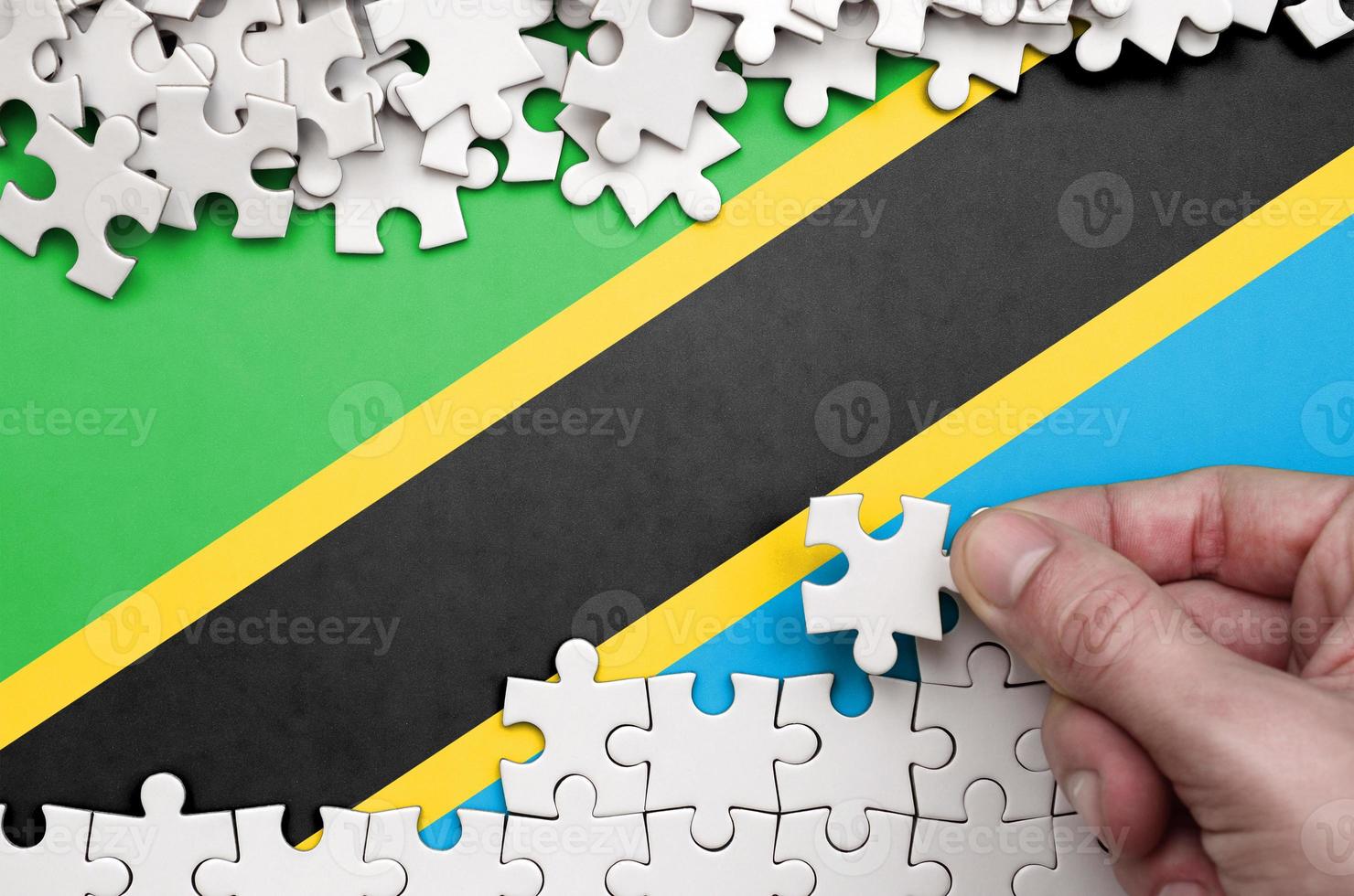 Tanzania flag is depicted on a table on which the human hand folds a puzzle of white color photo
