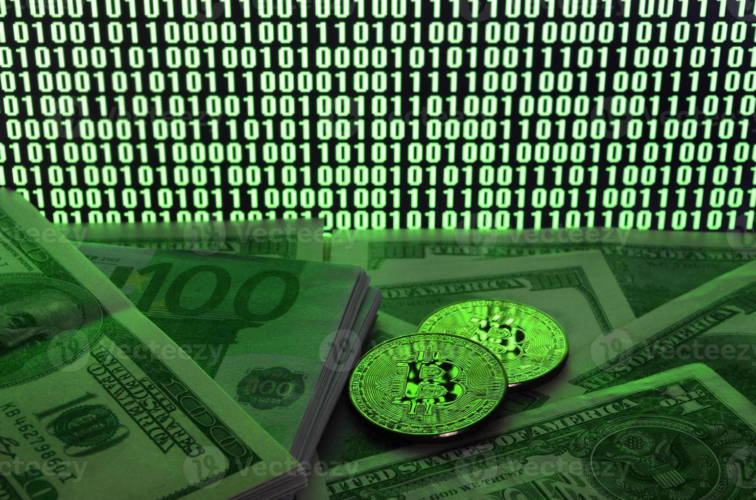 Two bitcoins lies on a pile of dollar bills on the background of a monitor depicting a binary code of bright green zeros and one units on a black background. Low key lighting photo