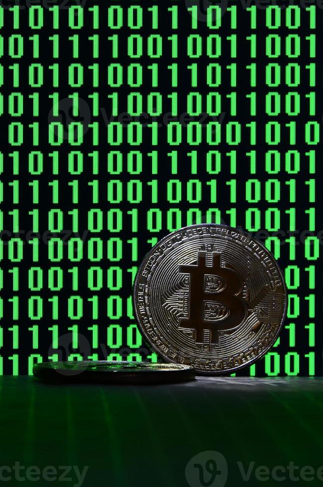 A pair of bitcoins lies on a cardboard surface on the background of a monitor depicting a binary code of bright green zeros and one units on a black background. Low key lighting photo