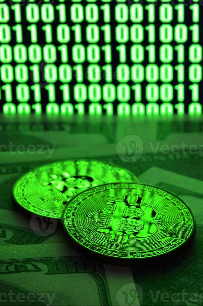 Two bitcoins lies on a pile of dollar bills on the background of a monitor depicting a binary code of bright green zeros and one units on a black background. Low key lighting photo