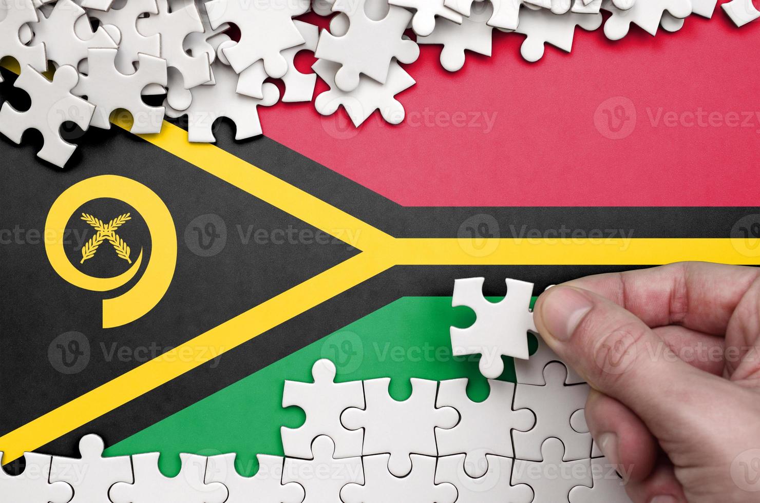 Vanuatu flag is depicted on a table on which the human hand folds a puzzle of white color photo