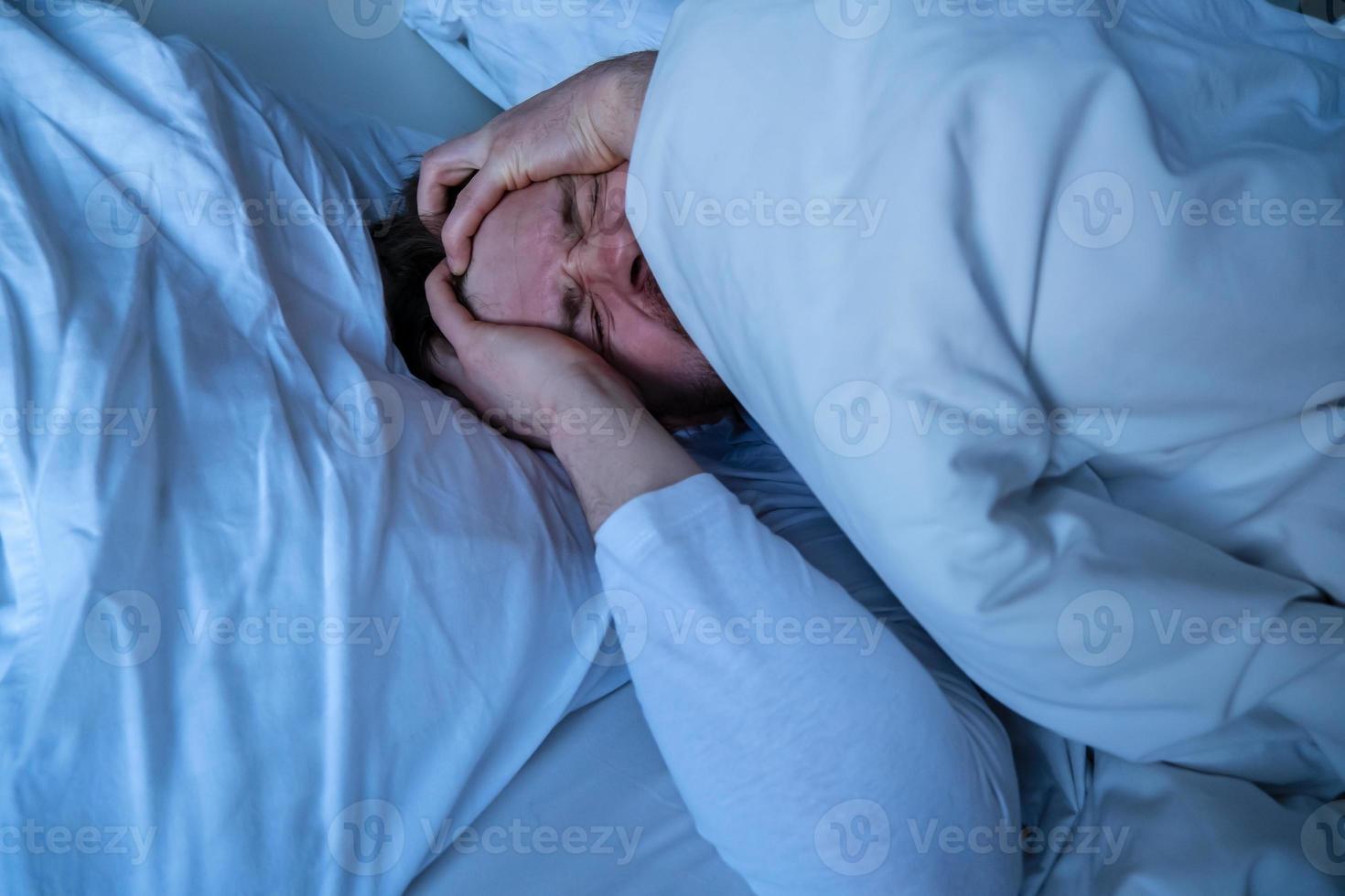Young man lies in bed with a severe headache, he clasps head with hands and makes a grimace of suffering. photo