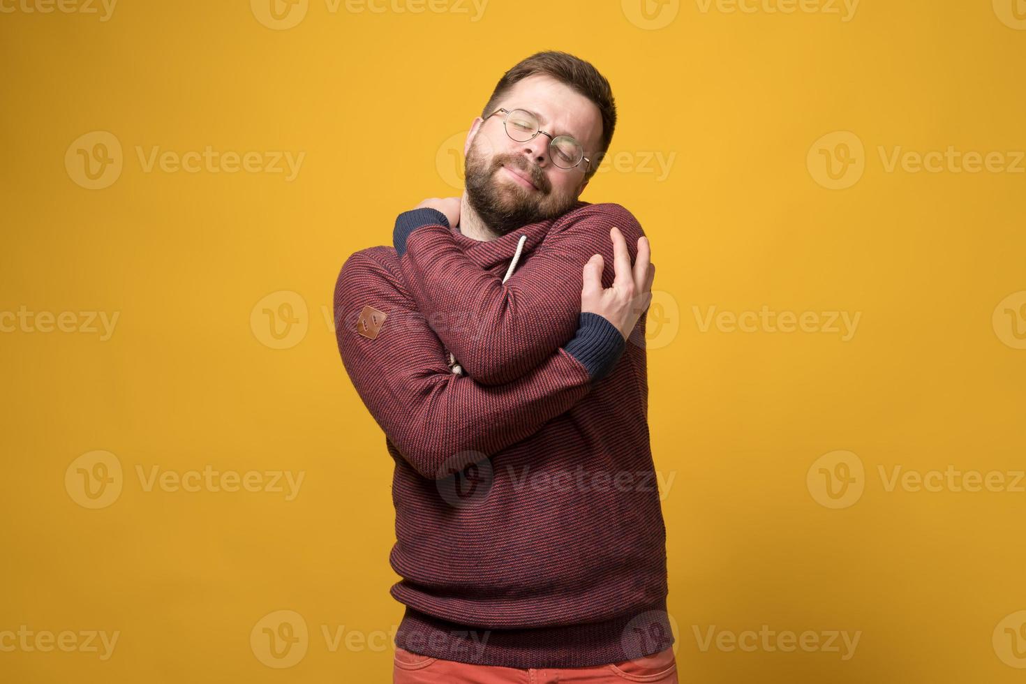 Charming Caucasian bearded man in glasses tenderly hugs himself, dreamily closing his eyes. Yellow background. photo