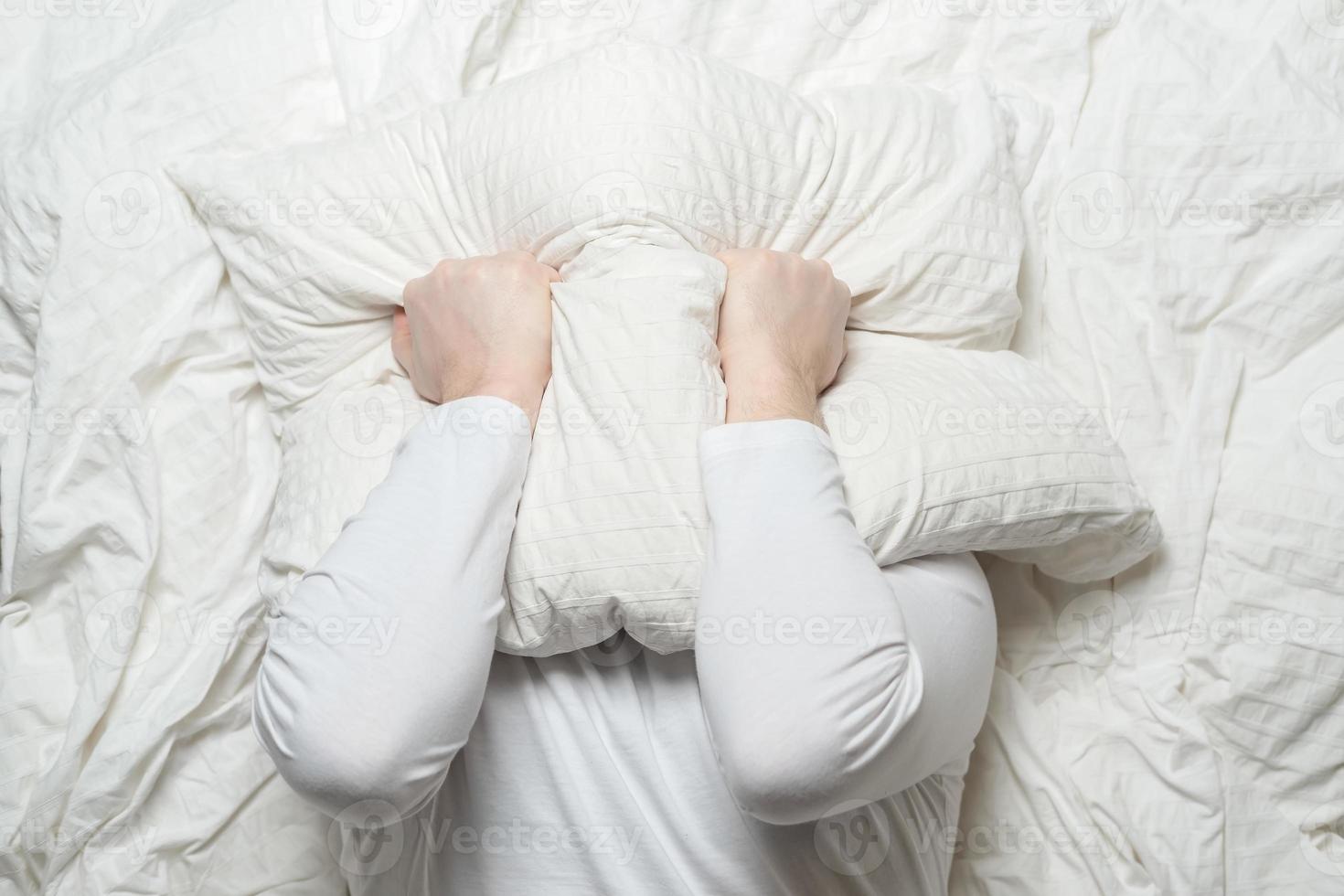 Man in white pajamas cannot sleep because of the noise and, in stress, covers head with a pillow. Insomnia concept. Top view. photo