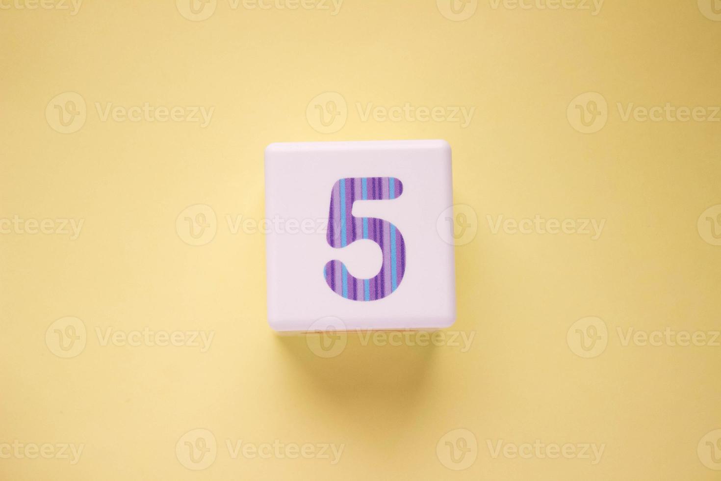 Close-up photo of a white plastic cube with number 5 on a yellow background. Object in the center of the photo