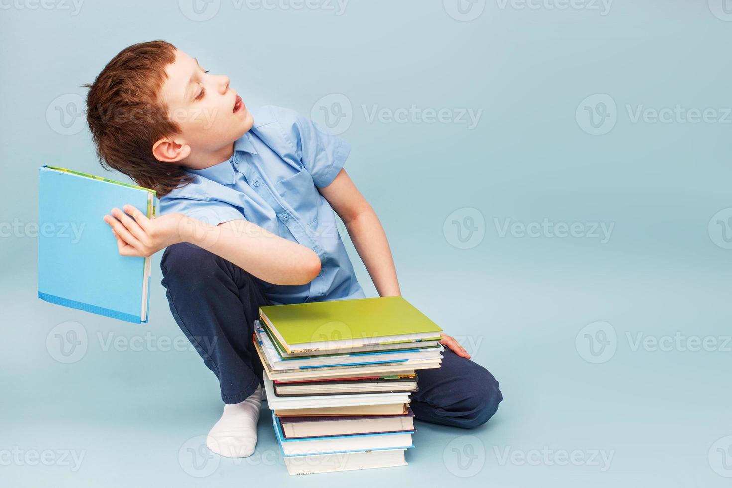 schoolboy sitting with pile of school books and throwing a textbook isolated on a blue background photo