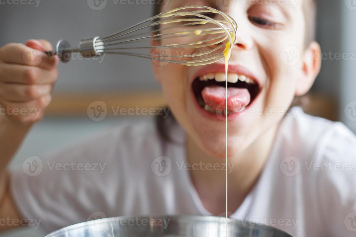 niño cocinando en la cocina. niño revolviendo masa para un pastel en el recipiente de acero. el niño prueba la comida y lame un batidor foto