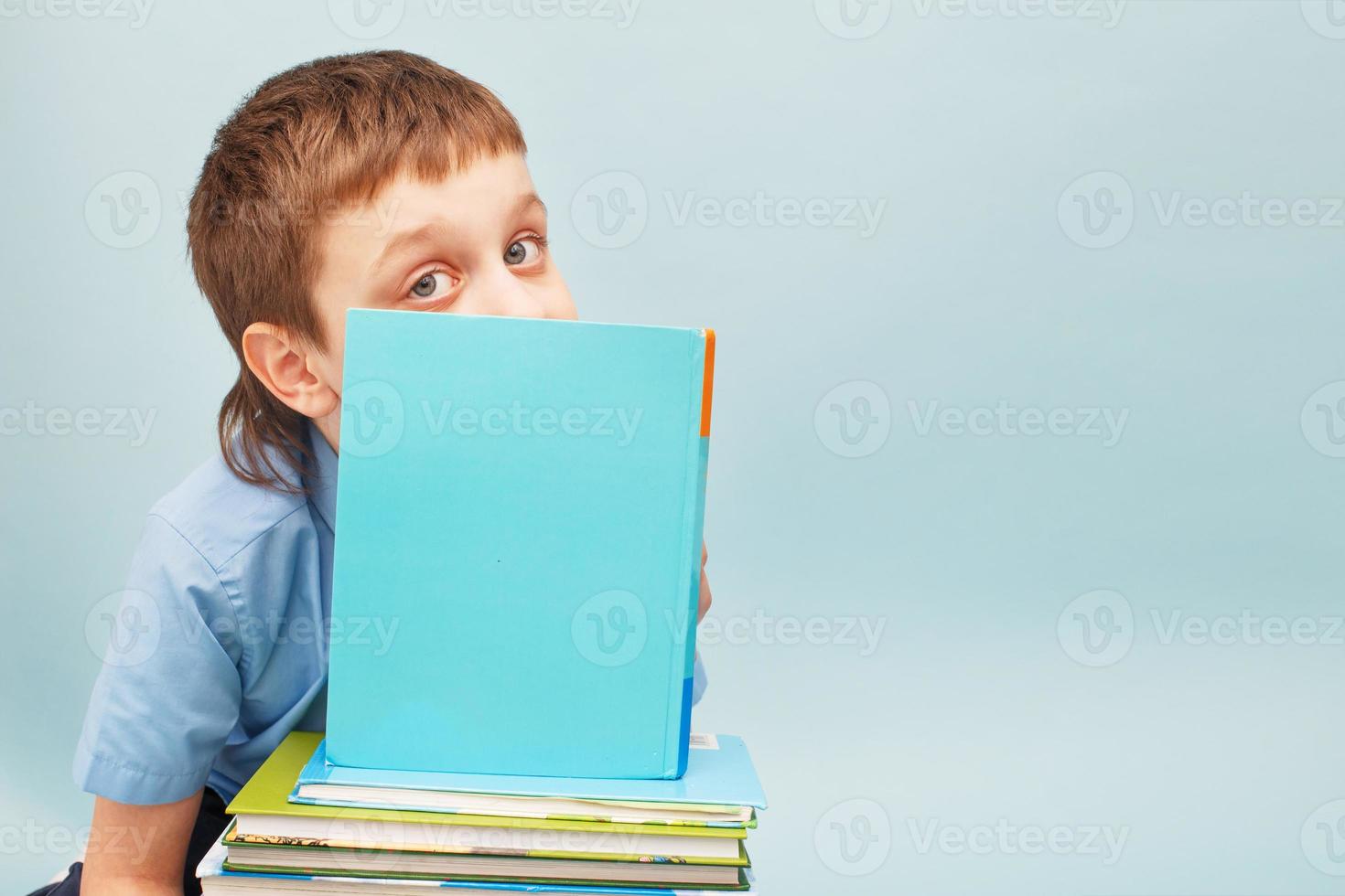 Schoolboy is sitting with a stack of books and reading and covers his face with a book isolated on blue background photo