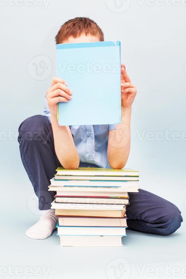 Schoolboy is sitting with a stack of books and reading and covers his face with a book isolated on blue background photo