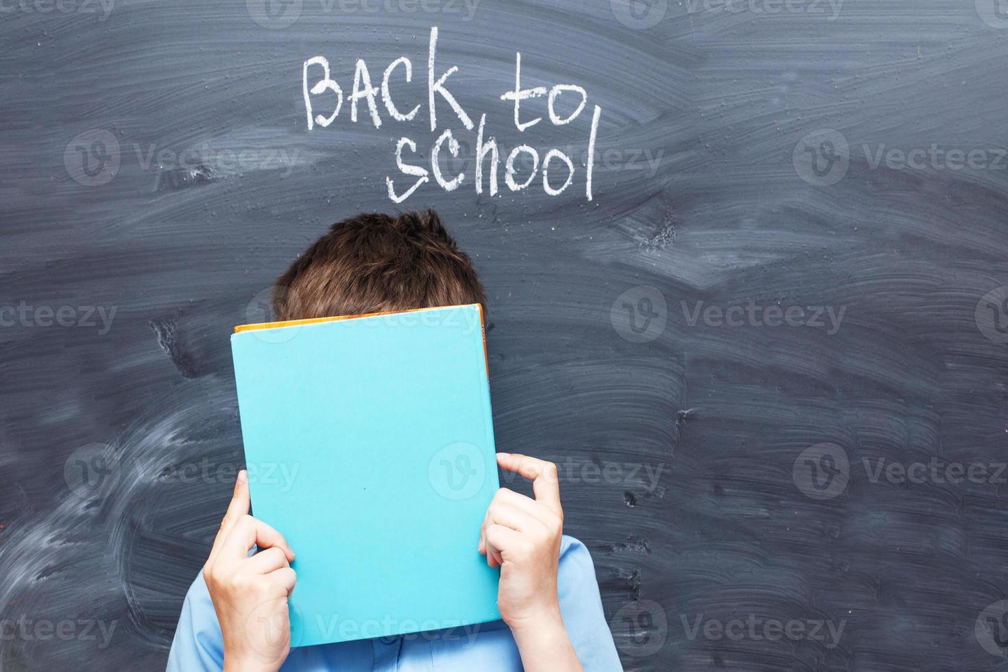 child boy covered his face with a textbook near chalkboard. Back to school concept background photo