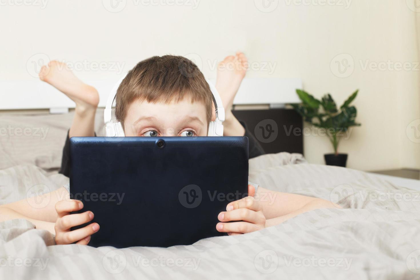 child boy lying on bed and covered his face with a pc tablet with wireless headphones photo