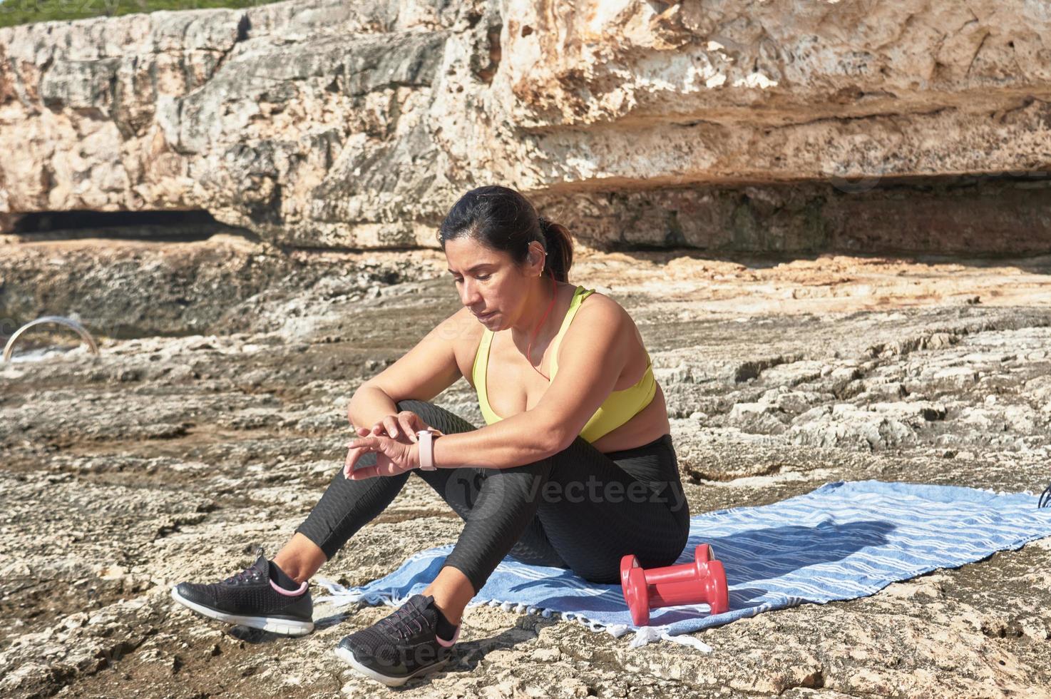 mujer latina, de mediana edad, mirando la aplicación, reloj inteligente, descansando después de la sesión de gimnasia, pesas, quemando calorías, manteniéndose en forma, al aire libre junto al mar, cielo azul, hermoso día foto