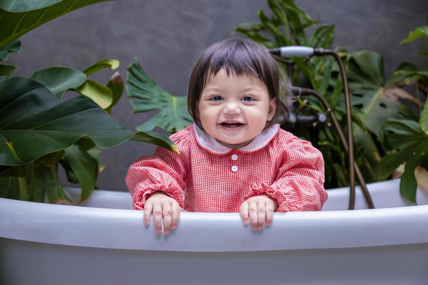 Caucasian little toddler baby girl smiling and playing inside the bathtub surrounding by tropical plant for better air purifying and sustainable home design concept photo