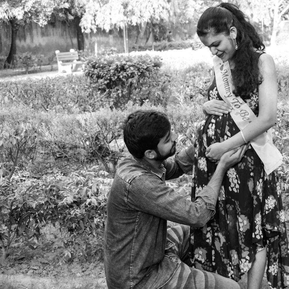 Indian couple posing for maternity baby shoot. The couple is posing in a lawn with green grass and the woman is falunting her baby bump in Lodhi Garden in New Delhi, India - Black and White photo