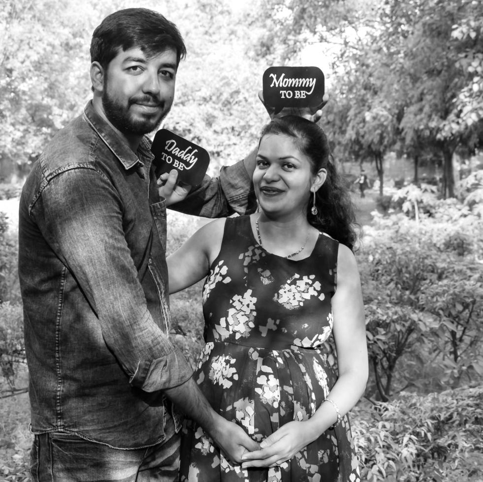 Indian couple posing for maternity baby shoot. The couple is posing in a lawn with green grass and the woman is falunting her baby bump in Lodhi Garden in New Delhi, India - Black and White photo