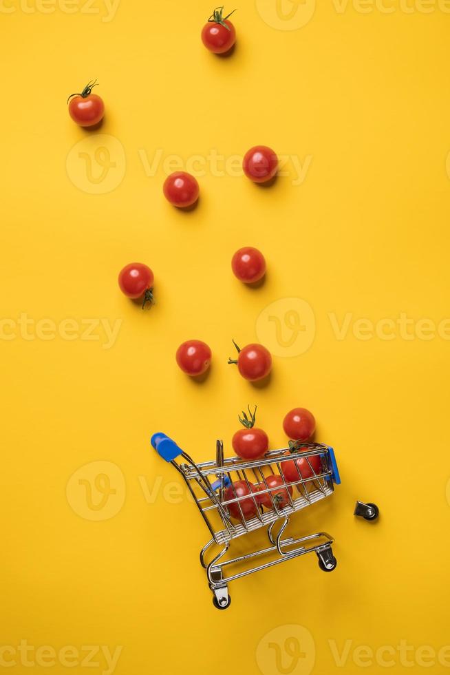 carro de supermercado decorativo volcado y tomates cherry que se cayeron de él, una rueda caída al lado, contra un fondo amarillo. vista superior. foto