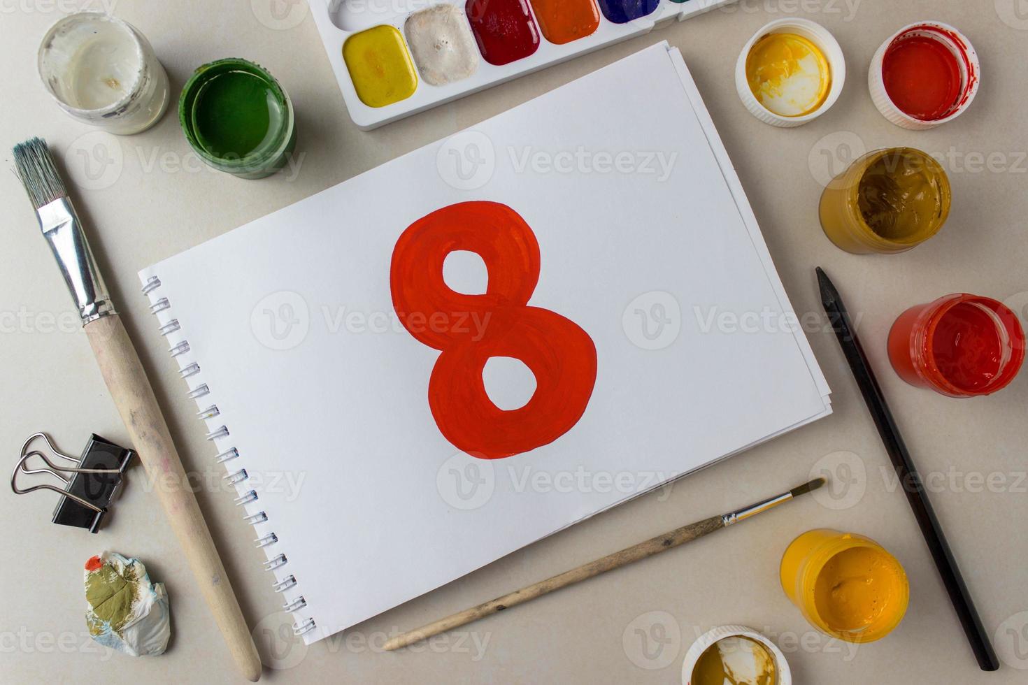 Red number eight drawing on a white album sheet. To indicate the place, price, etc. Nearby are brushes, jars of paint gouache, watercolor, etc photo