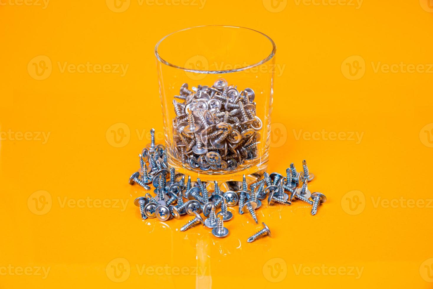 Many metal self-tapping screws made of steel in a glass, in bucket jar. self-tapping screw for metal, for iron, chrome-plated self-tapping screw, on an orange yellow background, photo