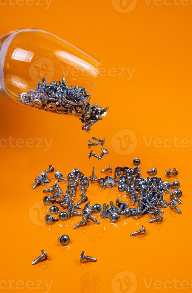 Dropped Many metal self-tapping screws made of steel in a glass, in bucket jar. self-tapping screw for metal, for iron, chrome-plated self-tapping screw, on an orange yellow background, photo