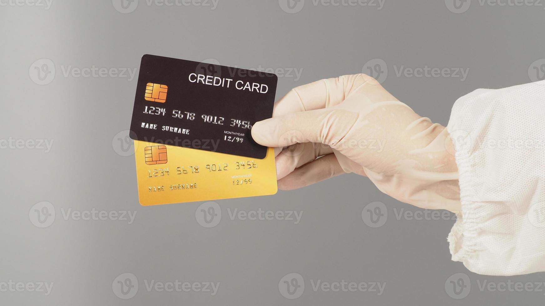 las manos sostienen dos tarjetas de crédito negras y doradas sobre fondo gris. la mano lleva un traje ppe y un guante médico blanco. foto