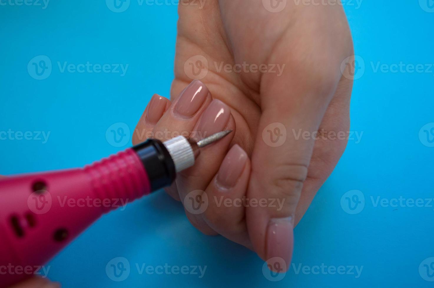 The process of making a beautiful manicure on the fingers of a finger processing a nail with a special machine in a nail beauty salon on a blue background photo