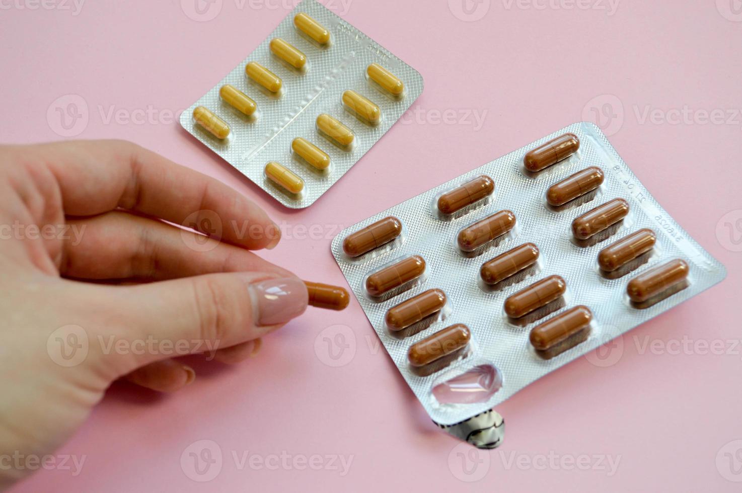 Oval brown and yellow tablets in individual packaging. capsule in the hands of a girl with manicure. taking pills for conception on a schedule. medicines on a pink background photo