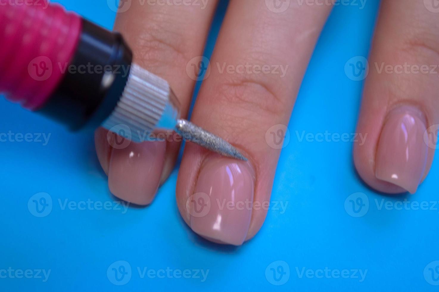 el proceso de hacer una hermosa manicura en los dedos de un dedo procesando una uña con una máquina especial en un salón de belleza de uñas sobre un fondo azul foto