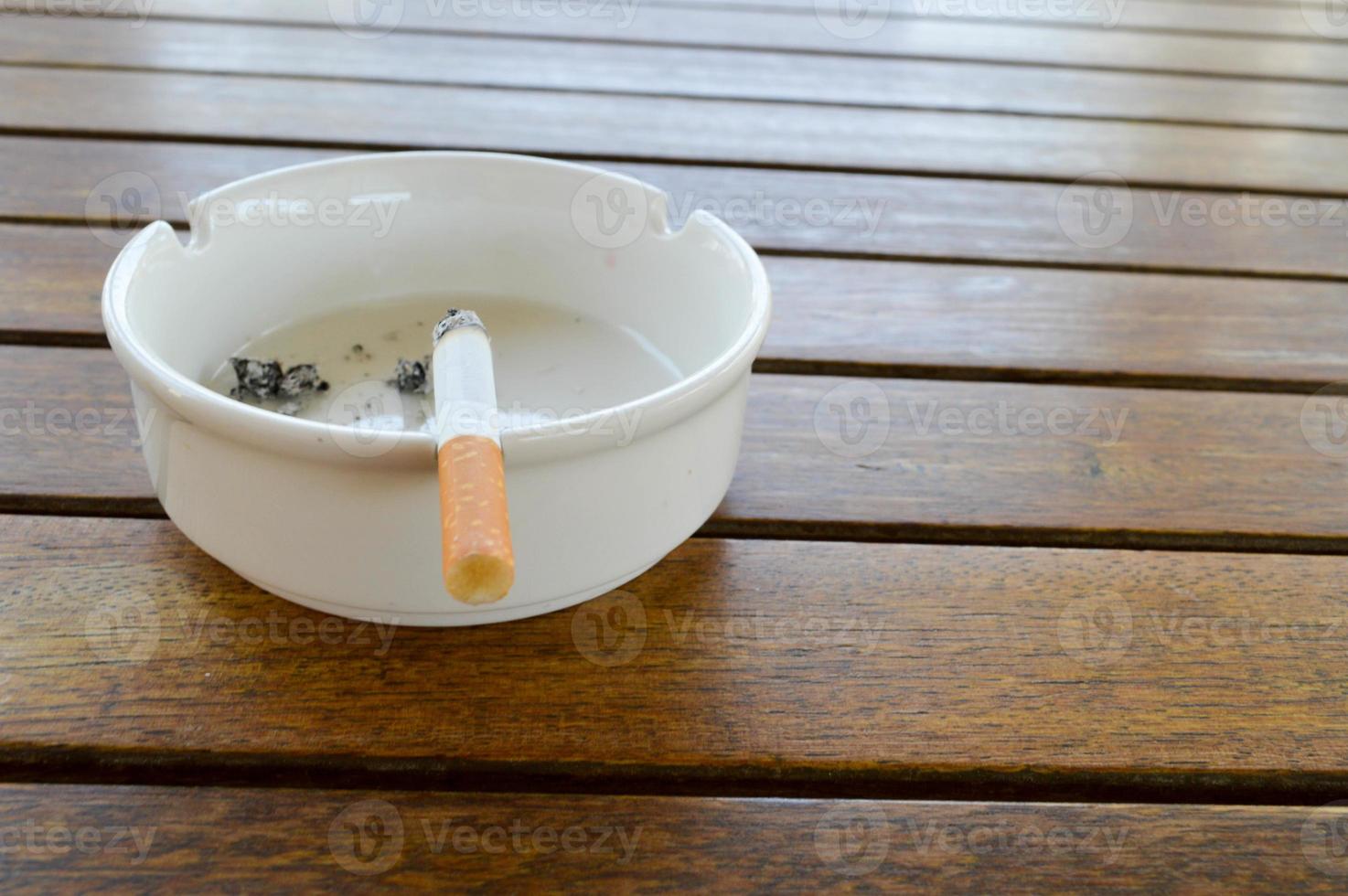 un cenicero de cerámica blanca con un cigarrillo y ceniza con un encendedor negro para fumar sobre una mesa de madera en un café foto