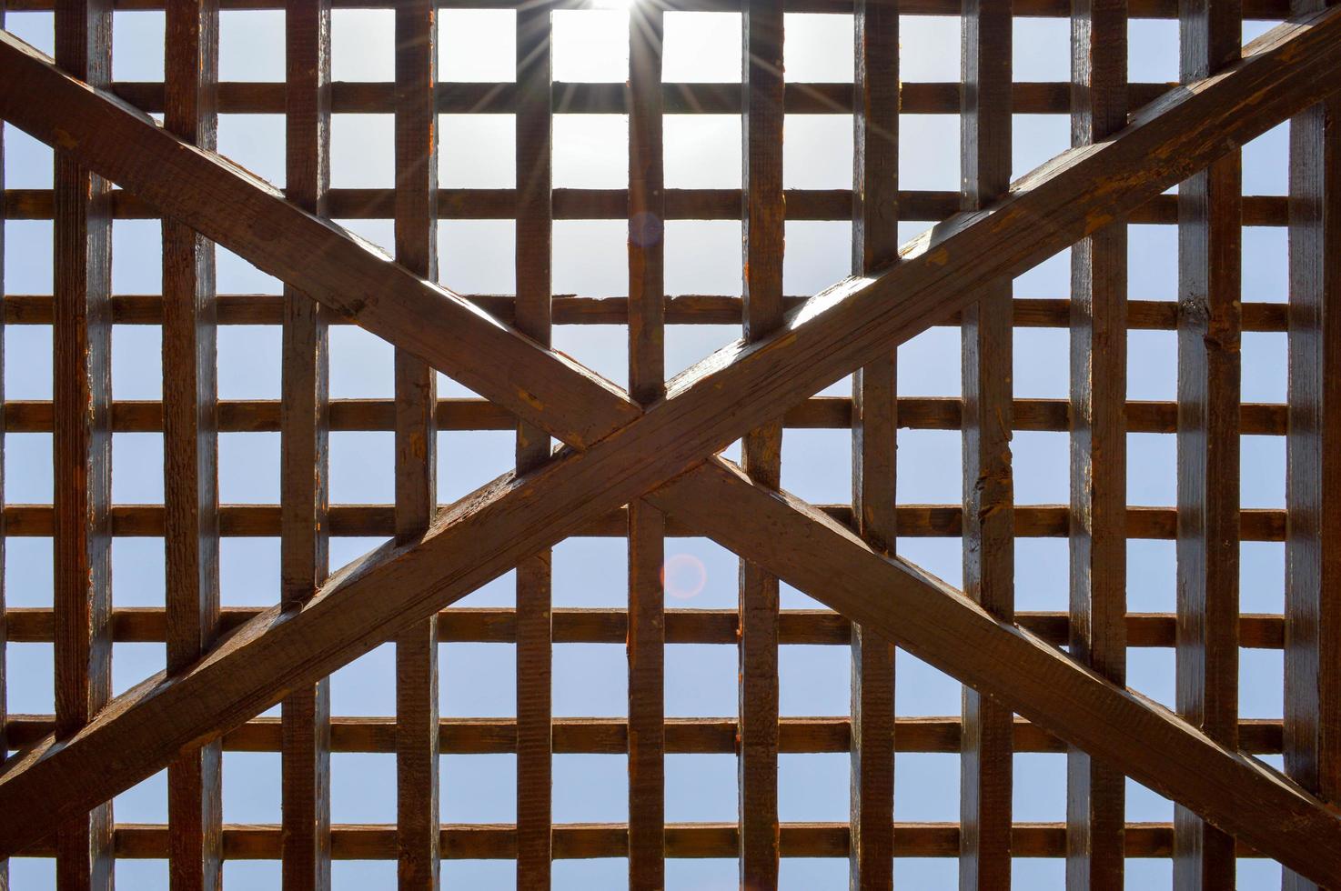 Texture of a brown wooden abstract lattice with square cells with holes from boards of log beams arranged vertically horizontally diagonally against the background of the sun and sky. The background photo