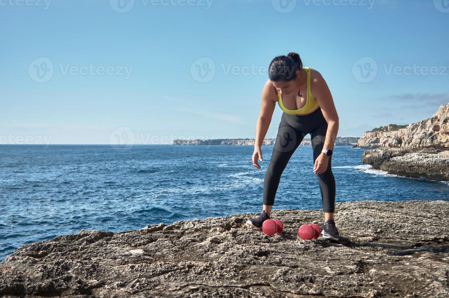 FITNESS LATIN WOMAN IN SPORTS SET TRAINING WITH ELASTIC BAND, WEIGHTS, GYM EXERCISES, IN FRONT OF THE WATER. photo