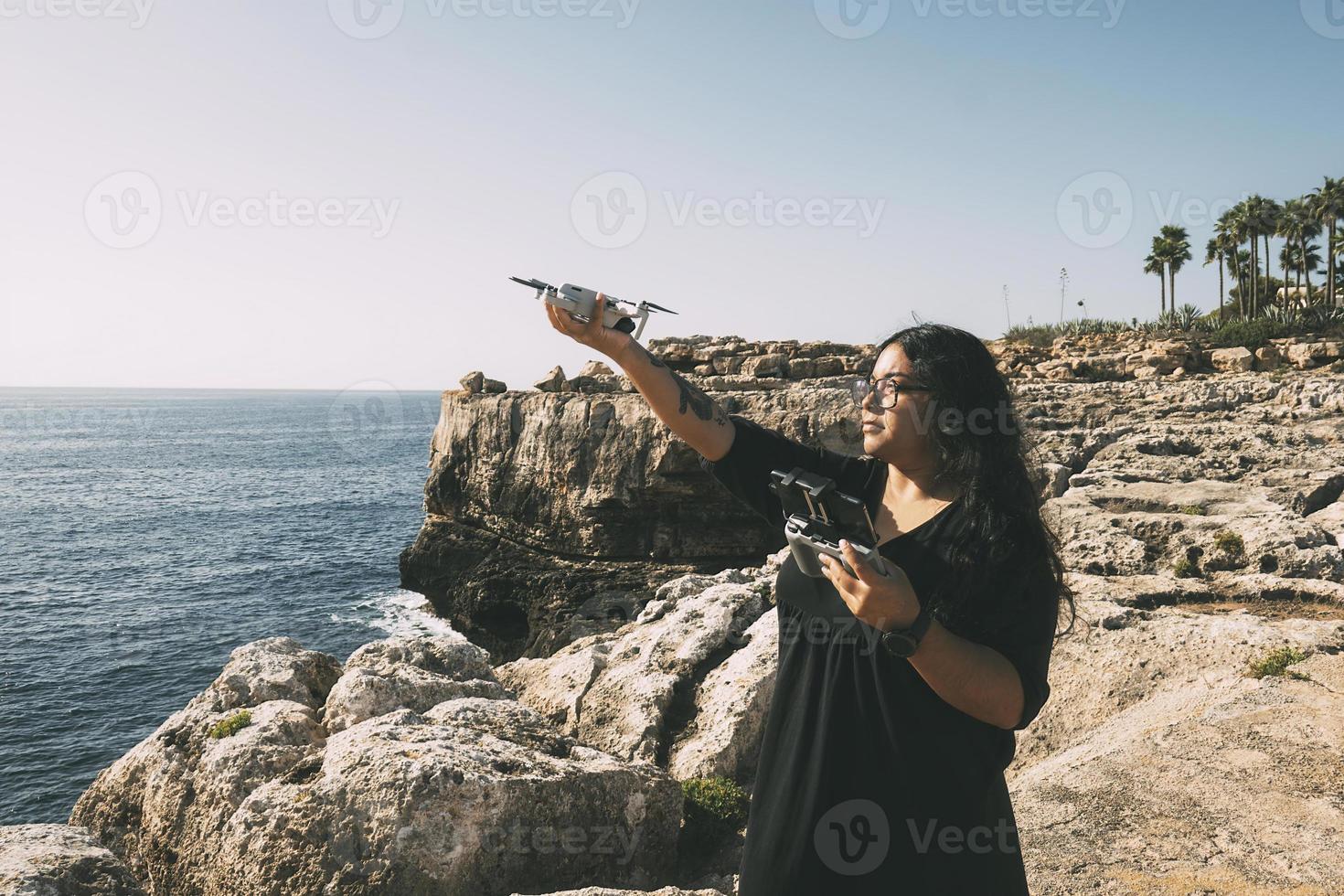 happy woman with drone, looking to the side black dress and remote control, on cliff, by the sea sunny day. technological concept photo