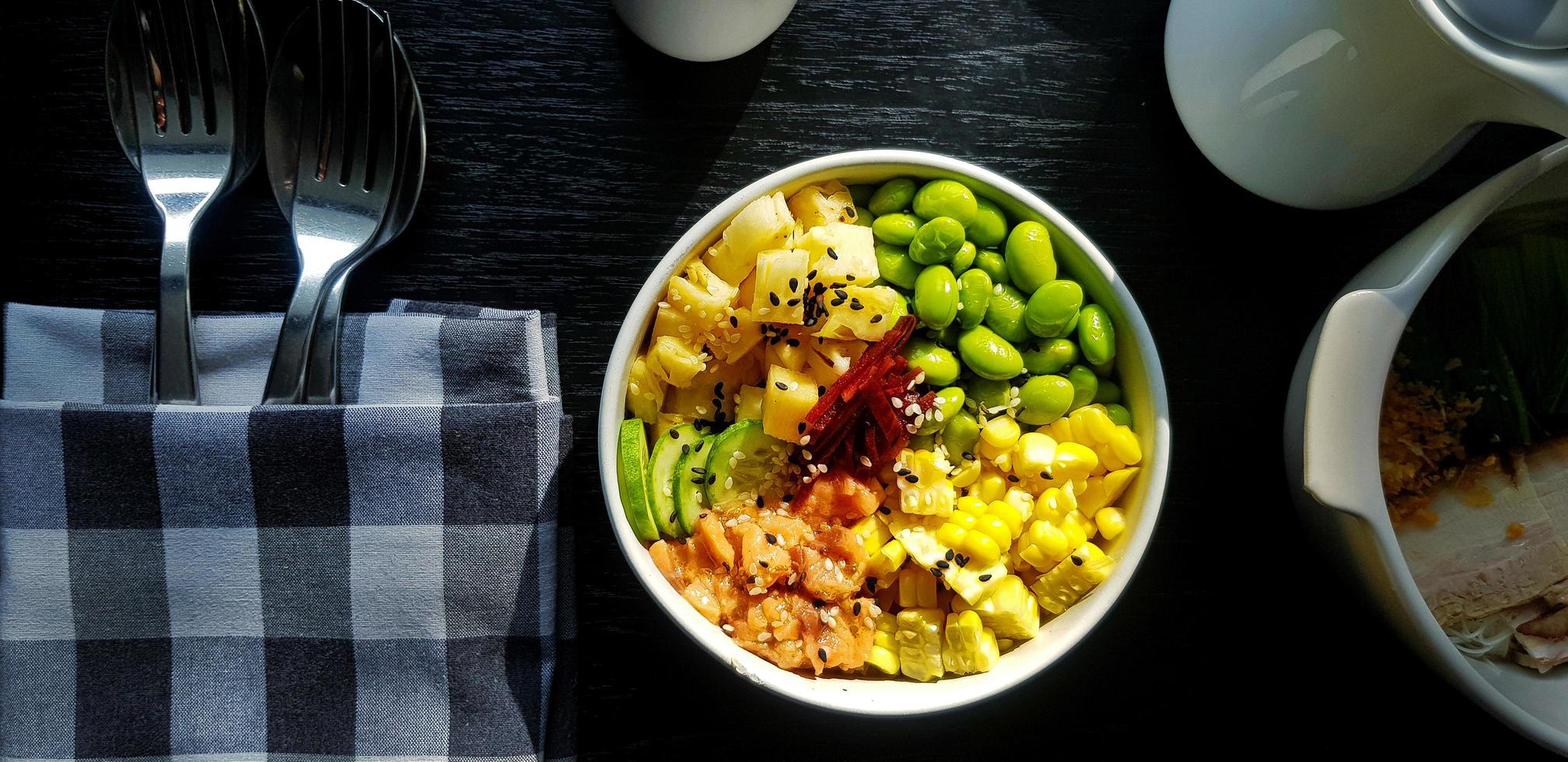 Endecha plana de ensalada fresca con rodajas de maíz, salmón, nuez verde o frijol, piña, tocino, pepino y cobertura con sésamo blanco y negro. conjunto de alimentos saludables con cuchara y tenedor en servilleta. foto