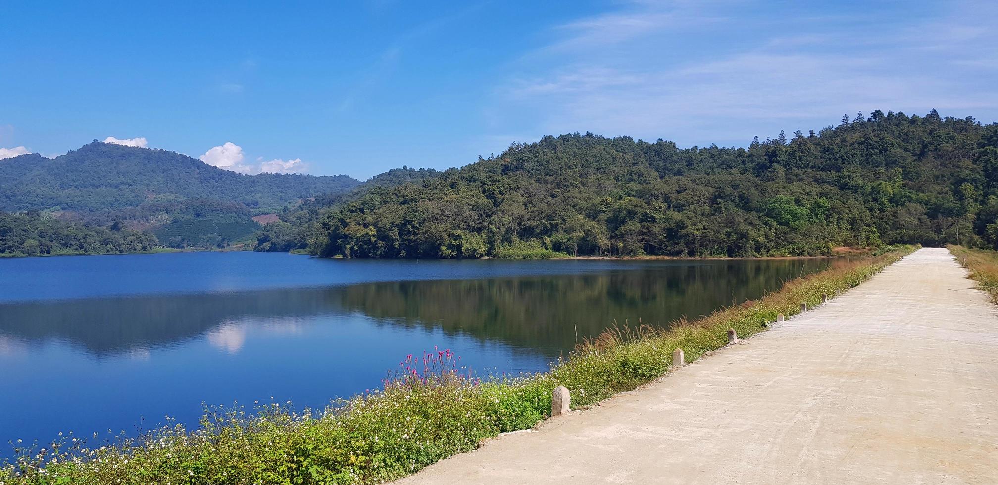 Road or street beside lake or river with green mountain and blue sky background at Huai Bon reservoir Chiang Mai, Thailand. Among nature with reflection on water and copy space. Beauty in nature photo