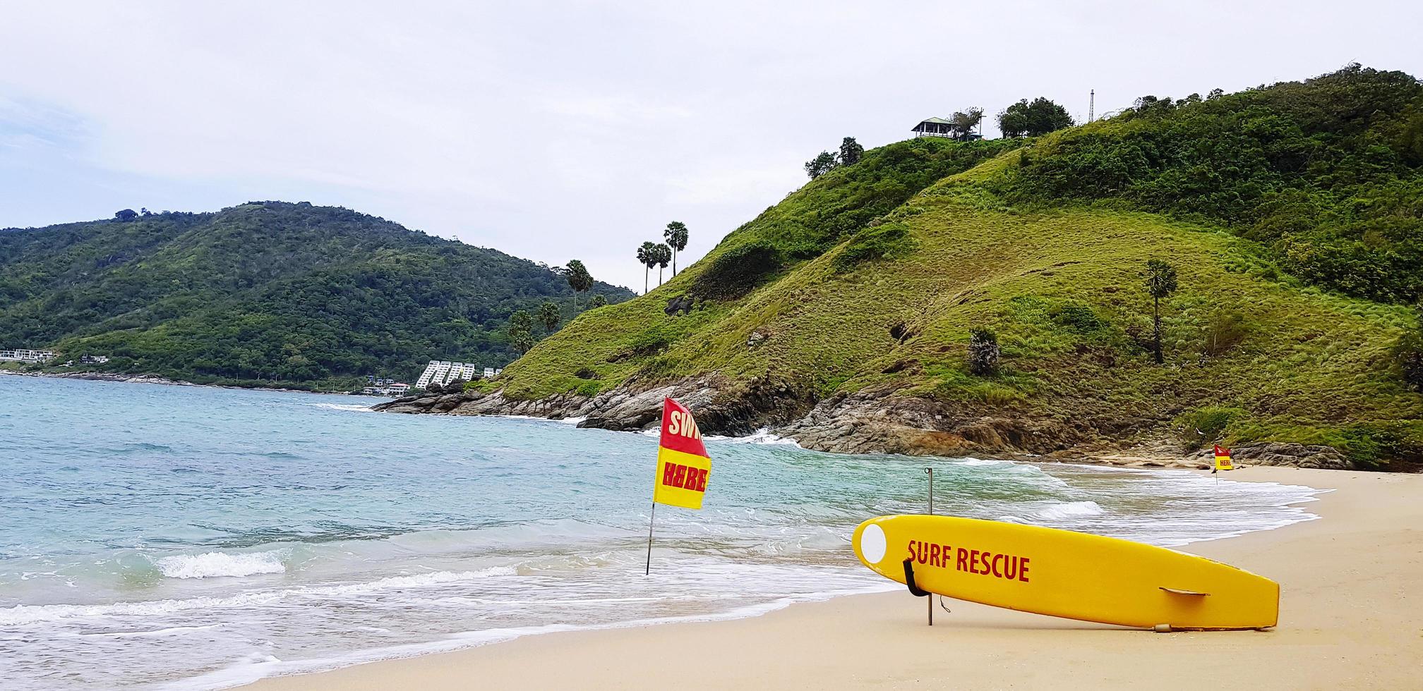 tabla de surf amarilla de rescate o guardia en la playa con bandera amarilla y roja para decirle a la gente o a los turistas que pueden nadar en el mar en esta área solo con fondo verde de montaña y cielo. belleza en la naturaleza con el océano. foto