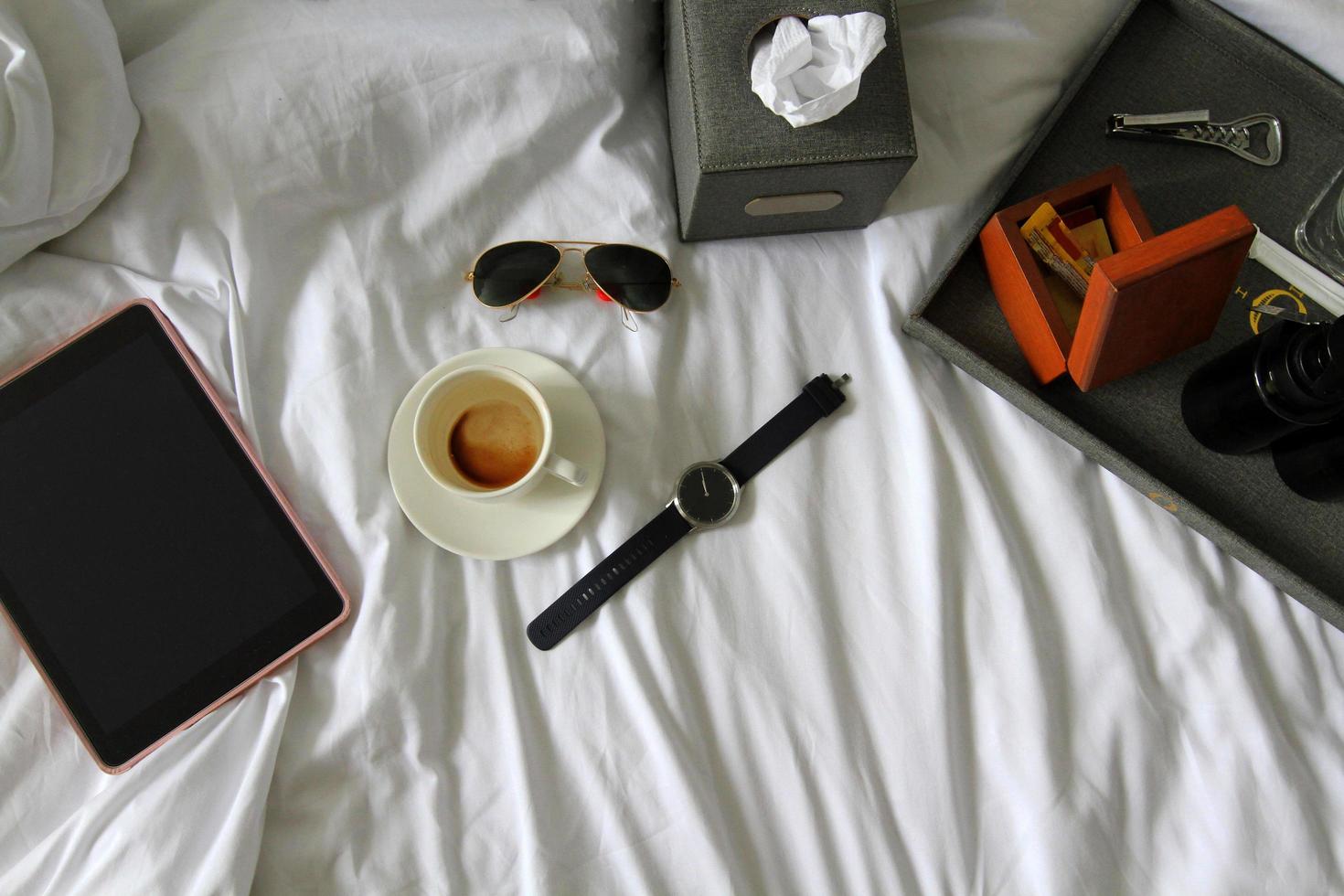 Top view of tablet, watch, black glasses, cup of coffee or tea, gray box of napkin , bottle opener, wooden box and plastic conditioner shampoo and bath shower gel on white bed. Workin, relax and drink photo