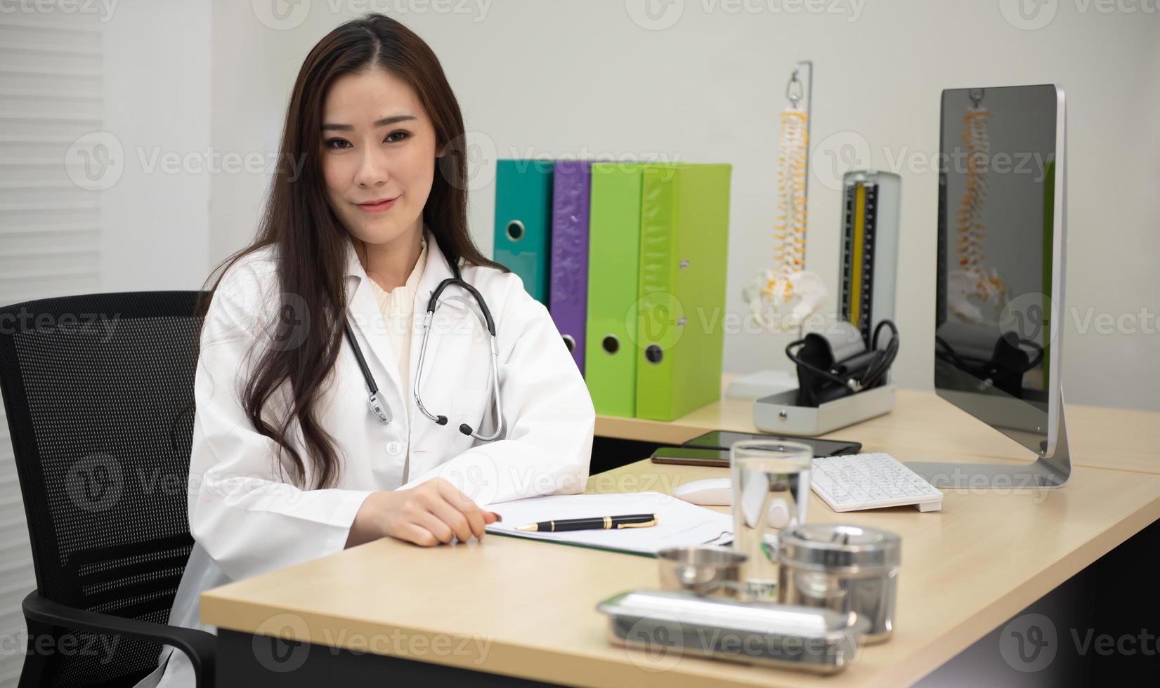 Portrait Of Smiling Asian Female Doctor Wearing White Coat With Stethoscope Sitting Behind Desk In Office. photo