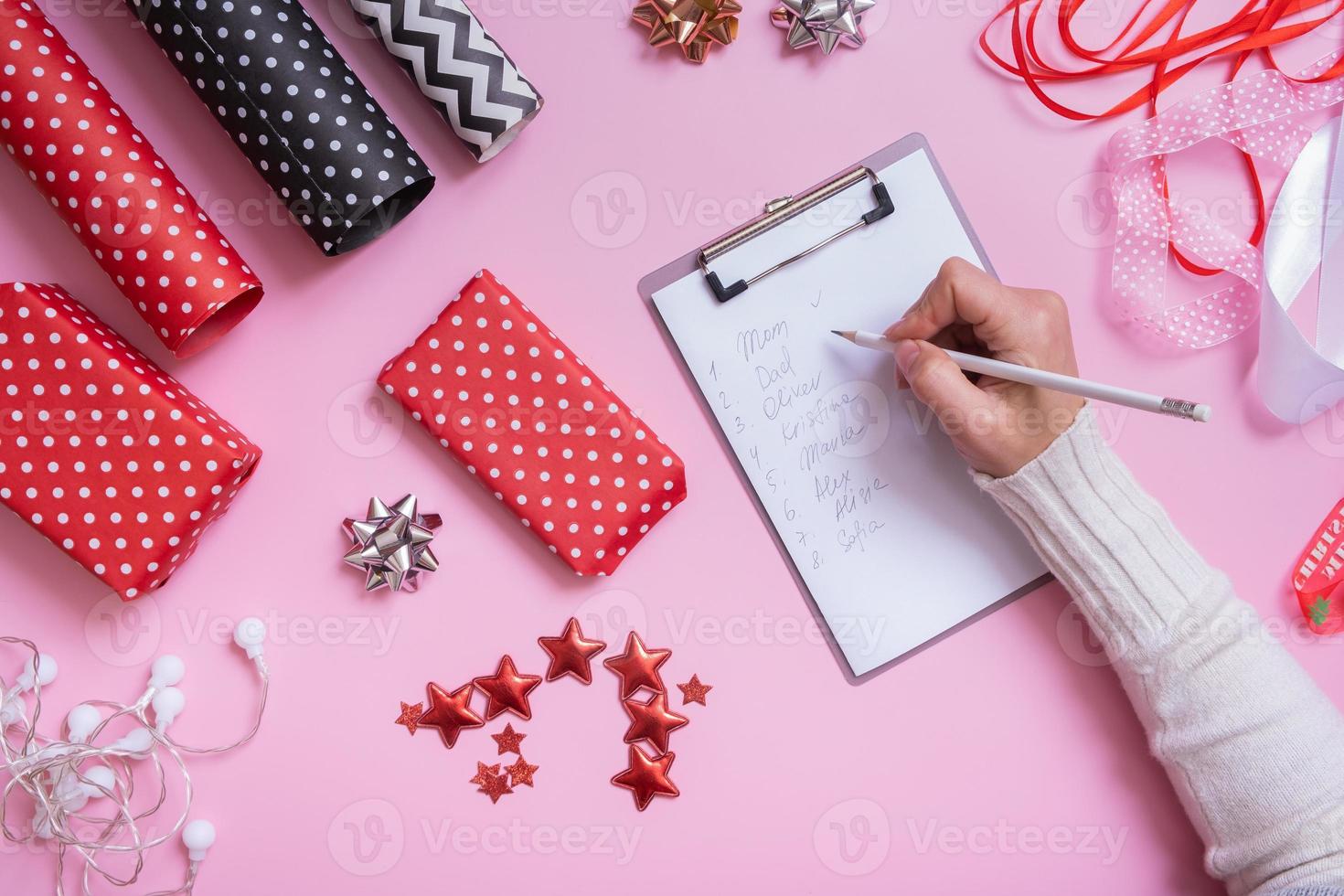 lista de nombres y envolver regalos de navidad planos sobre fondo rosa. vista superior del concepto de preparación navideña. foto