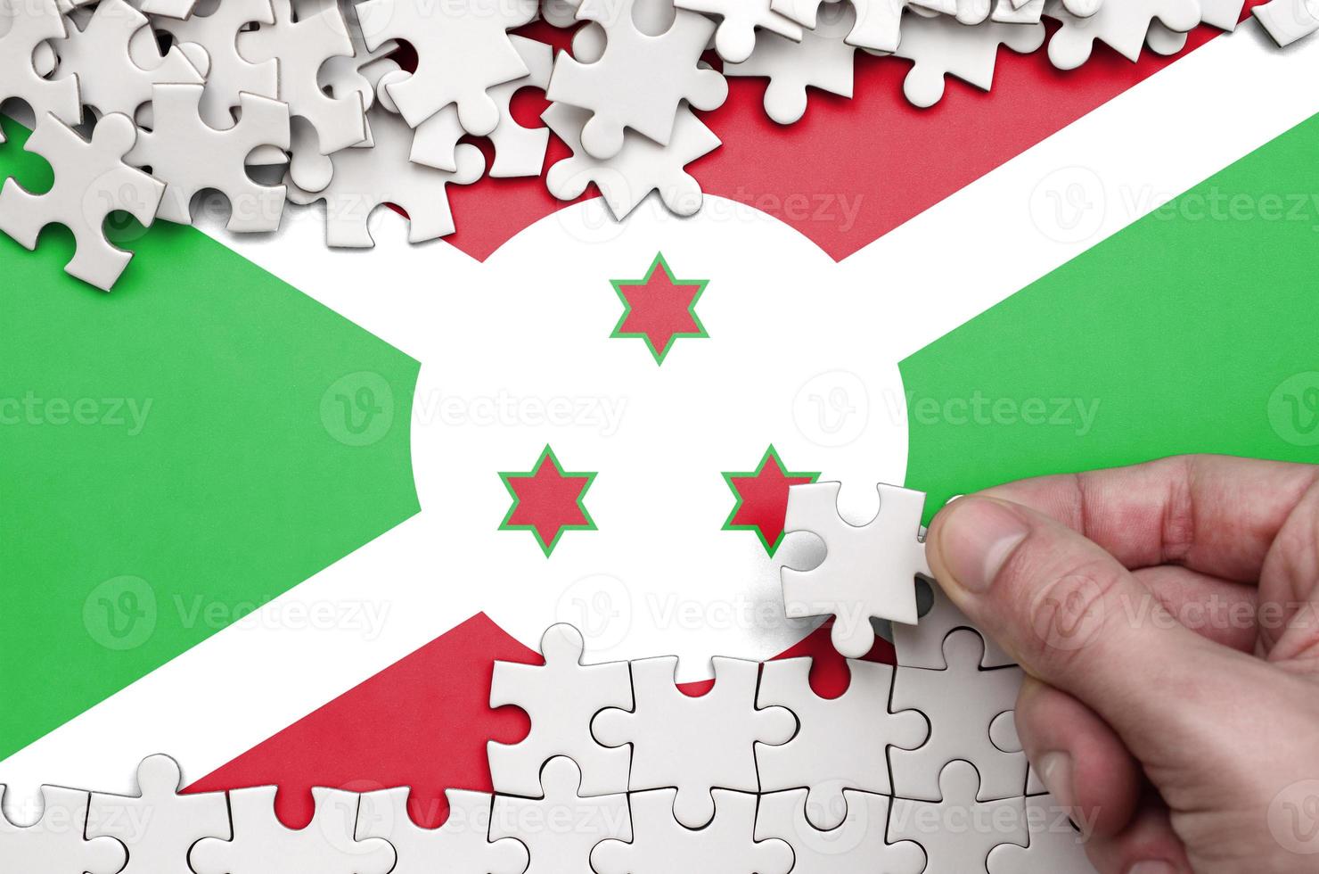Burundi flag  is depicted on a table on which the human hand folds a puzzle of white color photo
