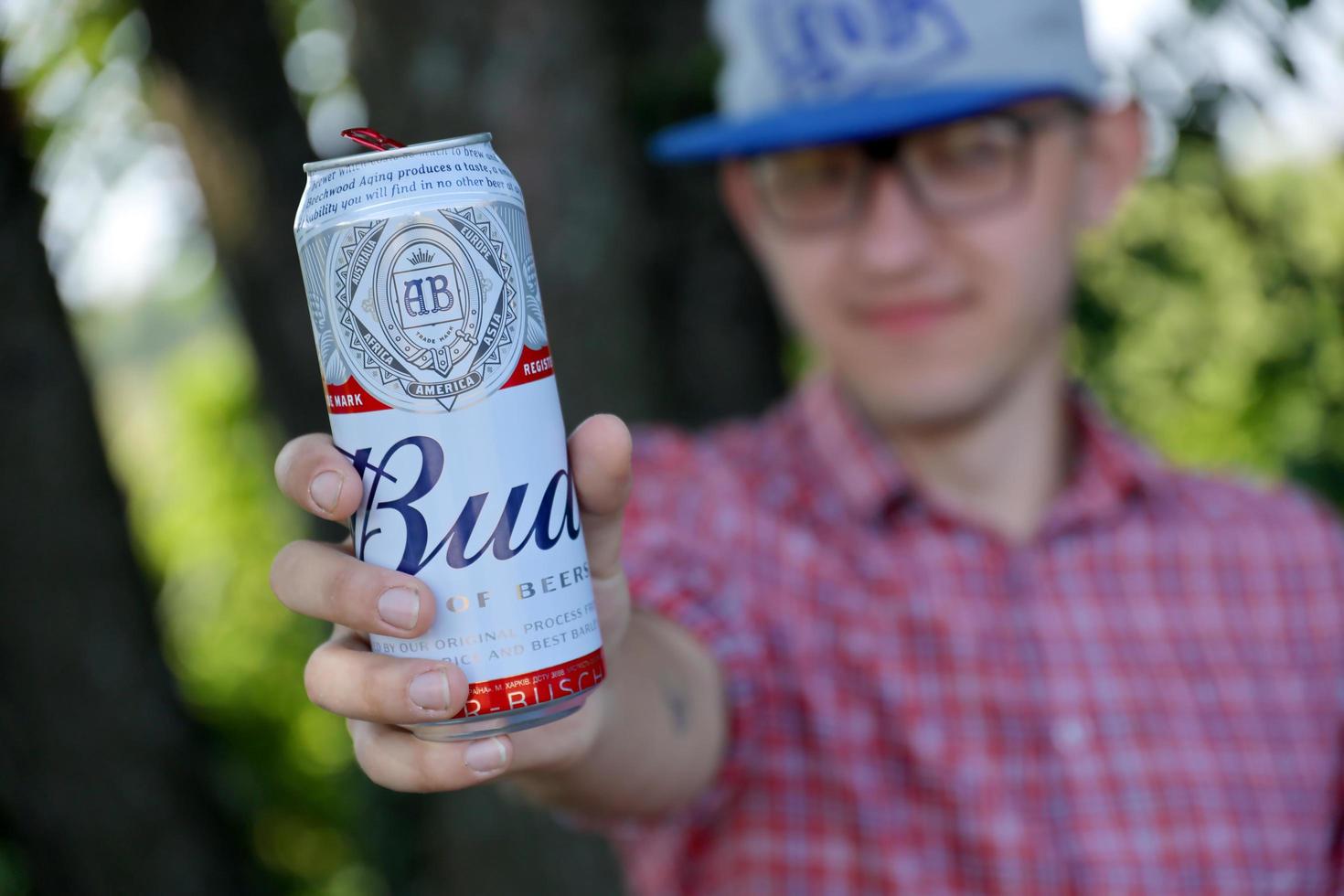 SUMY, UKRAINE - AUGUST 01, 2021 Young man raise Budweiser Bud beer can and shows BUD logo on blurred river and trees background. Budweiser is one of the most popular beer brands in the USA photo