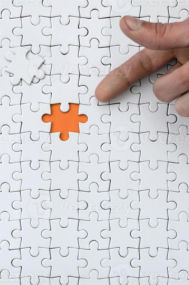 The texture of a white jigsaw puzzle in the assembled state with one missing element, forming an orange space, pointed to by the finger of the male hand photo