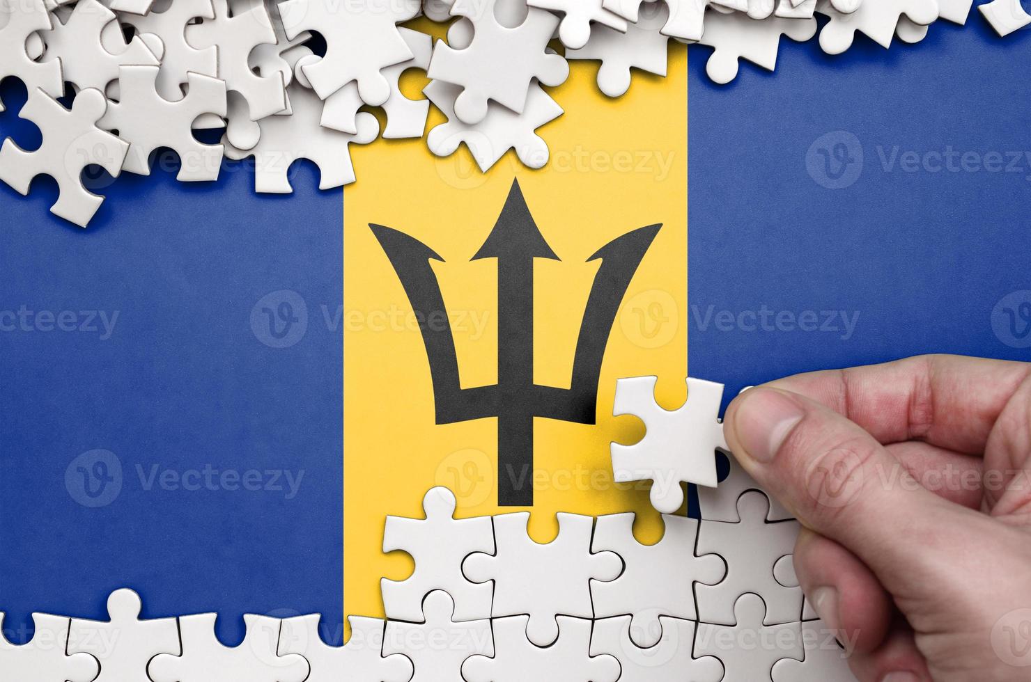 Barbados flag  is depicted on a table on which the human hand folds a puzzle of white color photo