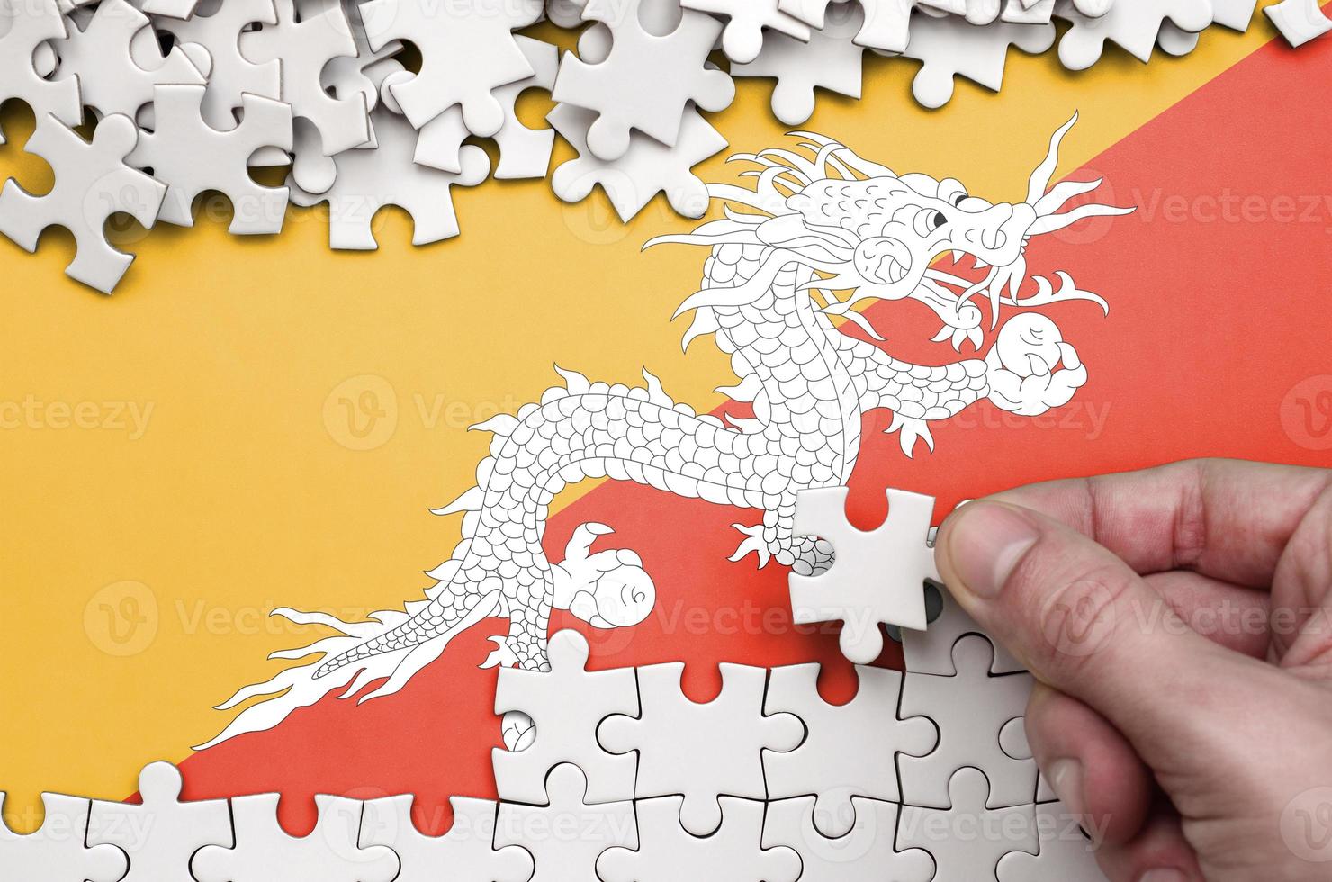Bhutan flag  is depicted on a table on which the human hand folds a puzzle of white color photo