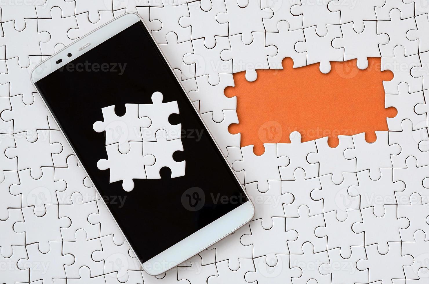 A modern big smartphone with several puzzle elements on the touch screen lies on a white jigsaw puzzle in an assembled state with missing elements photo