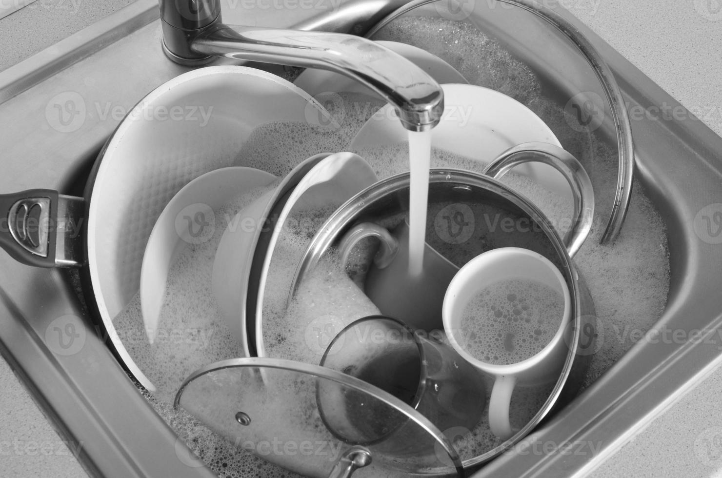 Dirty dishes and unwashed kitchen appliances lie in foam water under a tap from kitchen faucet photo