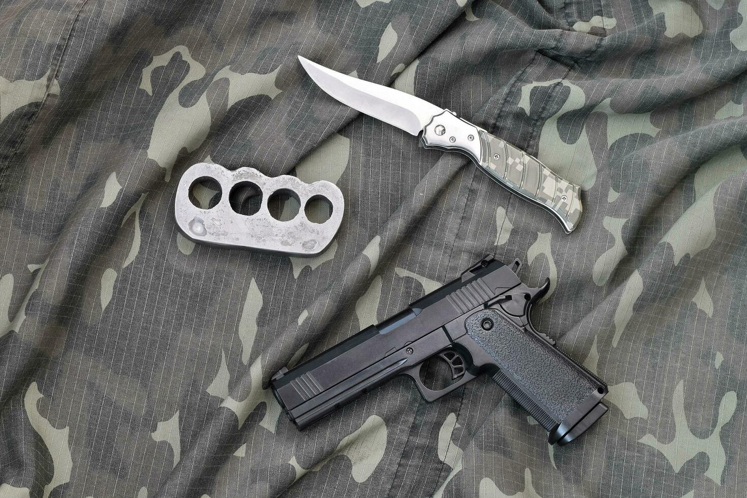 Handgun lies with brass knuckles and knife on camouflage military uniform close up. Concept of looting and arms trade photo