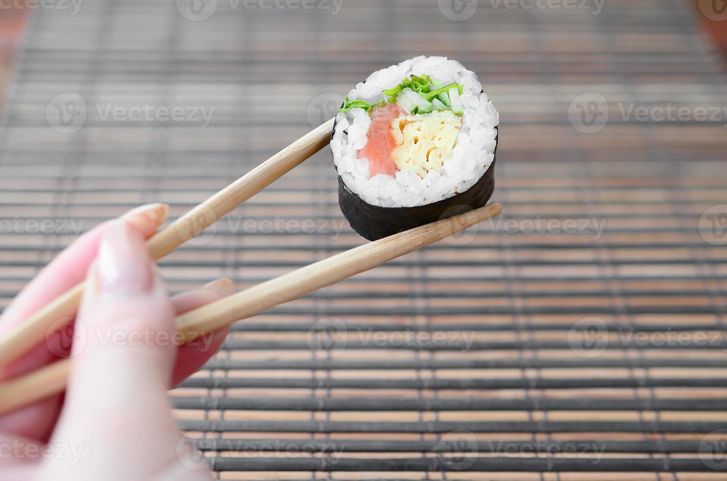 A hand with chopsticks holds a sushi roll on a bamboo straw serwing mat background. Traditional Asian food photo