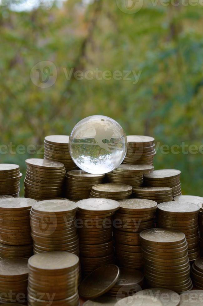 Glass planet on big pile of shiny ukrainian old 1 hryvnia coin stacks close up on blurred green trees background. The concept of money transfer around whole world photo